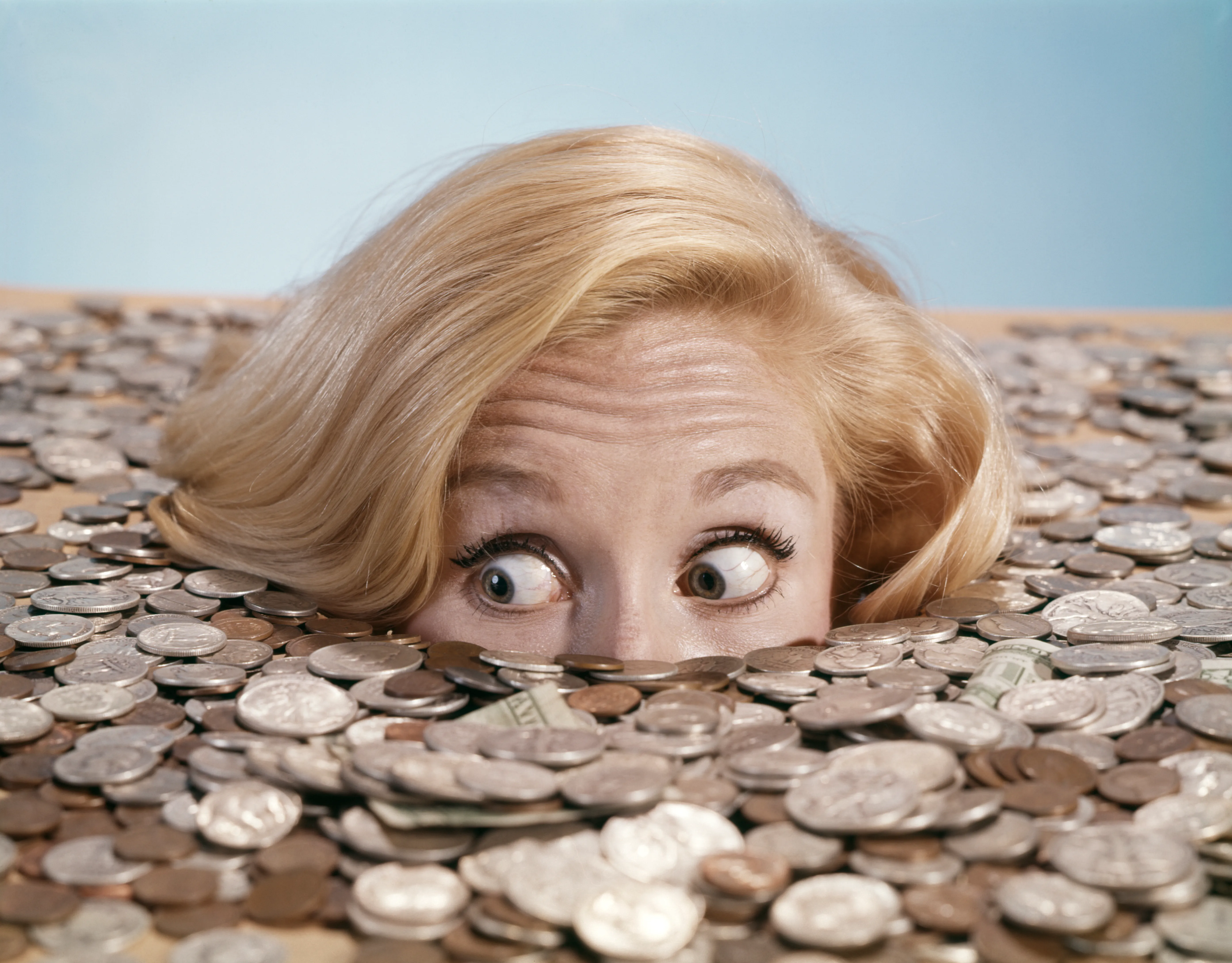 1960s blond woman buried up to her eyes in coins and bills looking alarmed