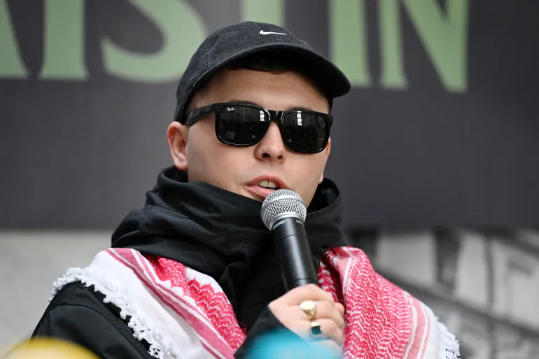 BELFAST, NORTHERN IRELAND - MARCH 11: Liam Óg Ó hAnnaidh, aka Mo Chara, of the band Kneecap speaks during a press conference following a High Court ruling which upheld the decision to drop the terrorism case against him on March 11, 2026 in Belfast, Northern Ireland. Irish language hip-hop group Kneecap called on supporters to attend the press conference in Belfast on Wednesday as the High Court in London ruled on the Crown Prosecution Service's (CPS) appeal on an earlier decision to throw out terror charges against rapper Liam Óg Ó hAnnaidh. Ó hAnnaidh, who performs with Kneecap under the stage name Mo Chara, was charged with a terror offence after allegedly displaying a flag in support of Hezbollah at a gig at the O2 Forum in Kentish Town in November 2024. The charge was dropped on a technicality in September 2025, which the CPS has appealed. (Photo by Charles McQuillan/Getty Images)