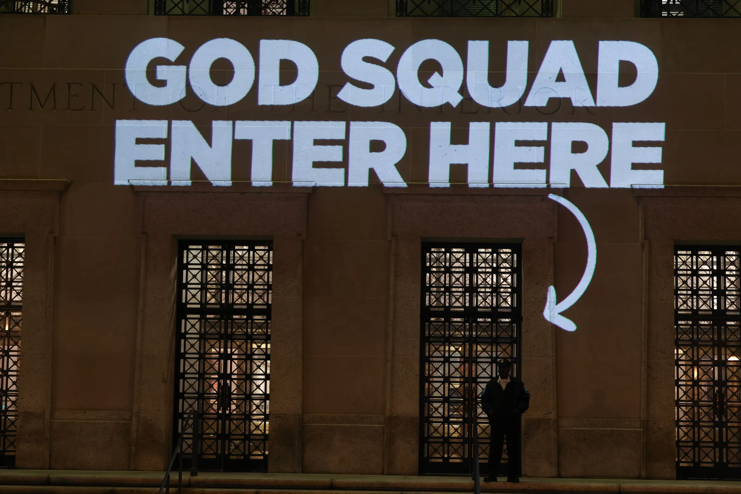 A guard stands outside the Department of Interior, with the words God squad enter here light up on the facade