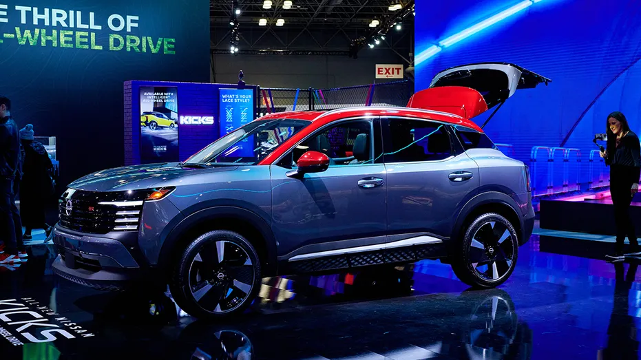 A gray and red Nissan Kicks compact SUV is displayed on an auto show floor under blue and purple lighting, with promotional signage and attendees visible in the background.