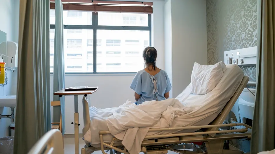 Female patient sitting on hospital bed wearing hospital gown in ward
