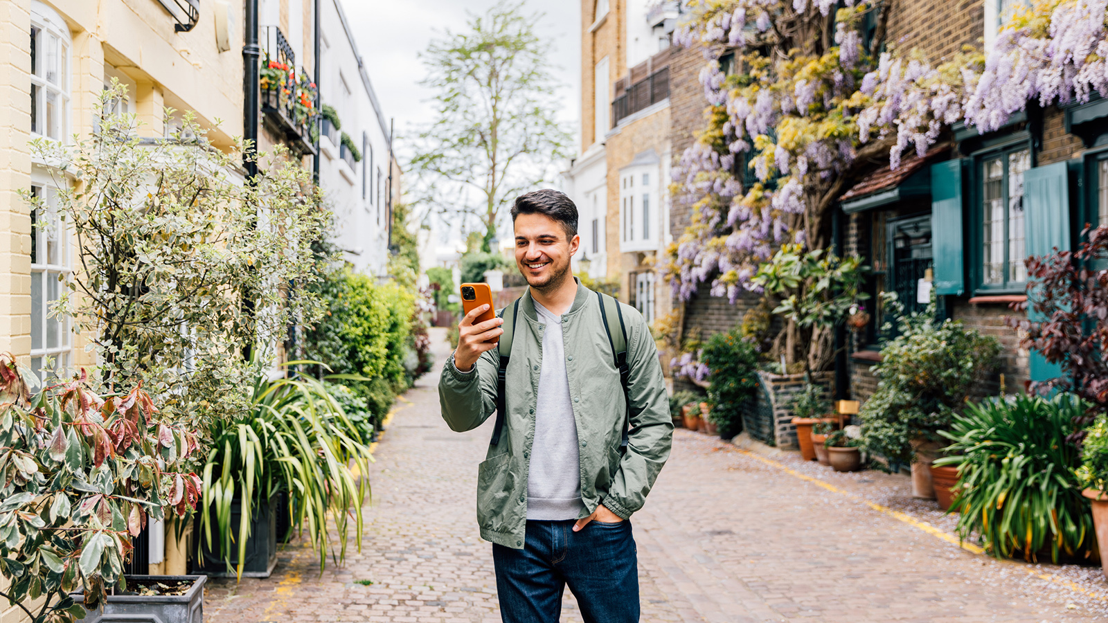 Young man smiling at phone while on a street lined with potted plants