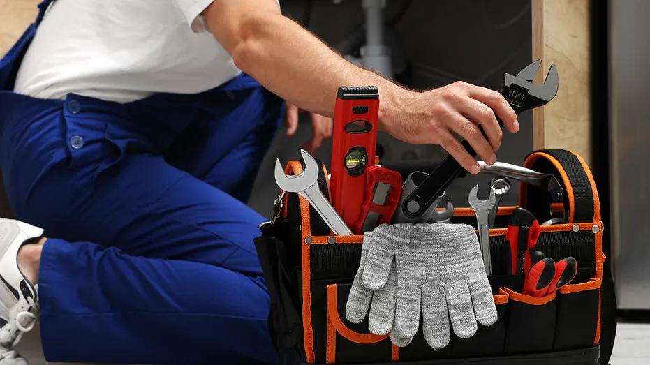 A plumber reaches for a wrench beside a tool bag filled with equipment.