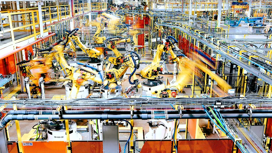 Multiple yellow robotic arms operate along an automotive manufacturing line inside a Ford factory.