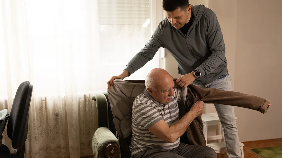 Caring middle aged son helping his senior father to get dressed while sitting in armchair at home